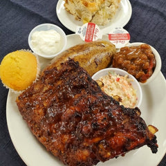 A dinner size plate filled with a half rack of ribs, coleslaw, baked potato, baked beans, cornbread, butter, sour cream and a dessert